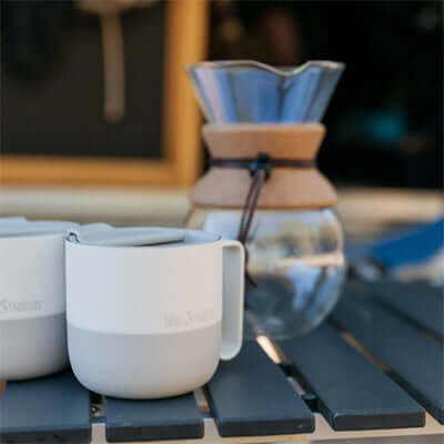 Klean Kanteen Rise 14oz Mug (with Flip Lid) on a table with a coffee maker in the background, perfect corporate gift option.