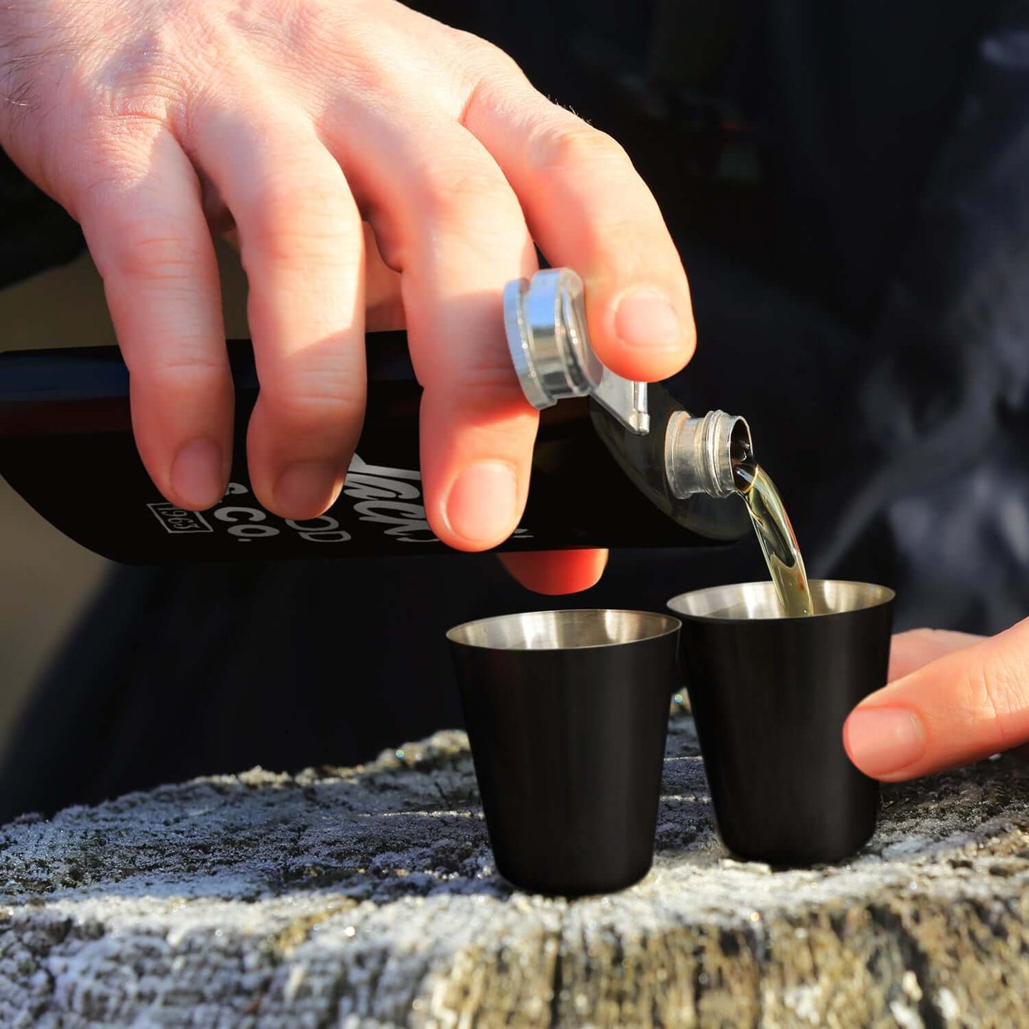 Person pouring whiskey from Dalmore Hip Flask Gift Set into two shot glasses outdoors.