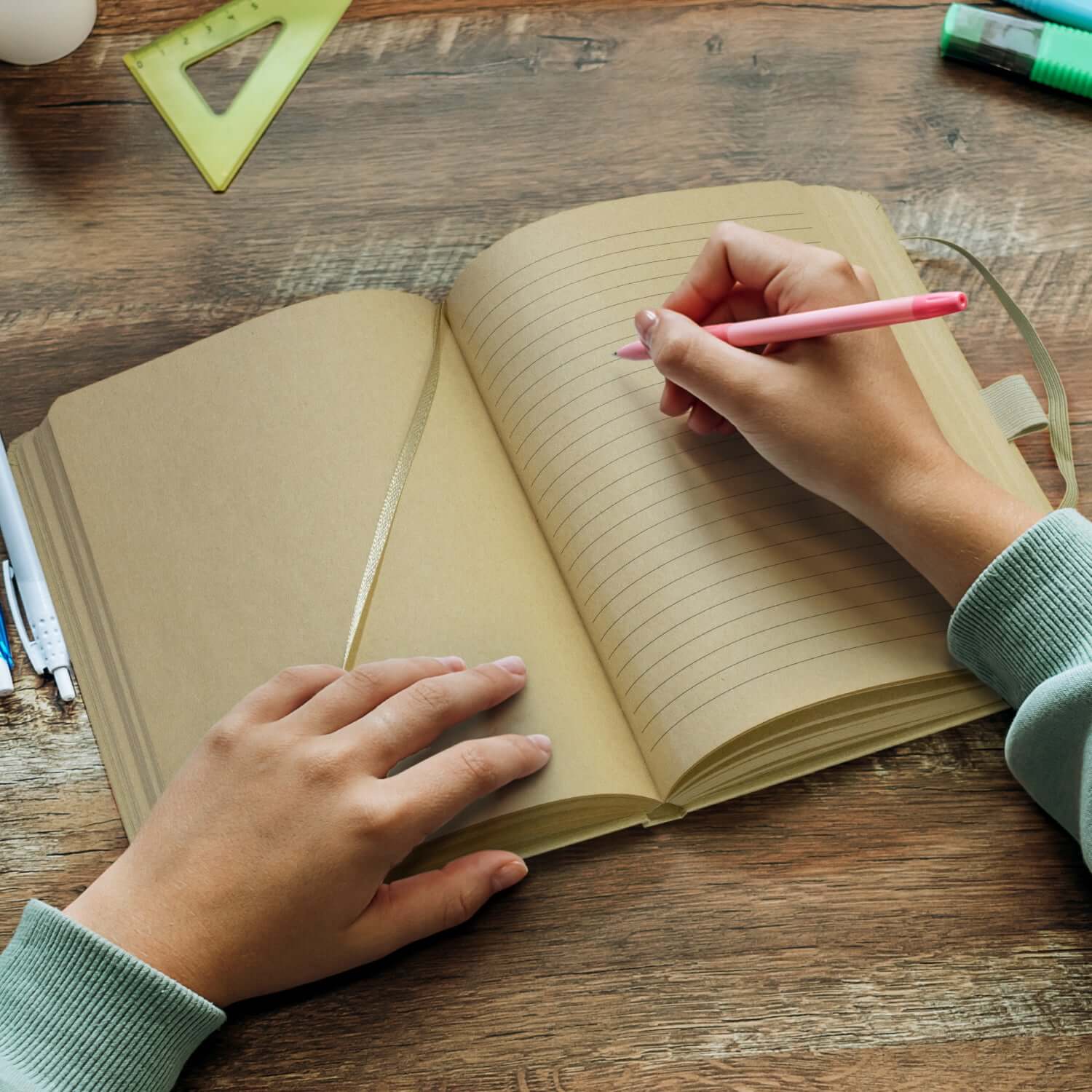 Person writing in the Kraft Omega Notebook with pink pen and ruler on a wooden desk.