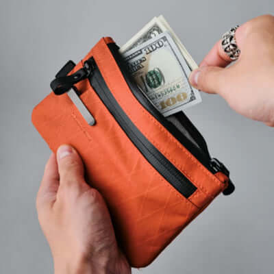 Person holding Alpaka Zip Clutch Cayenne RX30 + Hand Carry Strap with cash partially inside pocket.