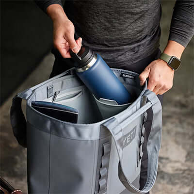 Person placing a Rambler bottle inside a Yeti Camino® 35 Carryall Tote Bag, showcasing its spacious design.