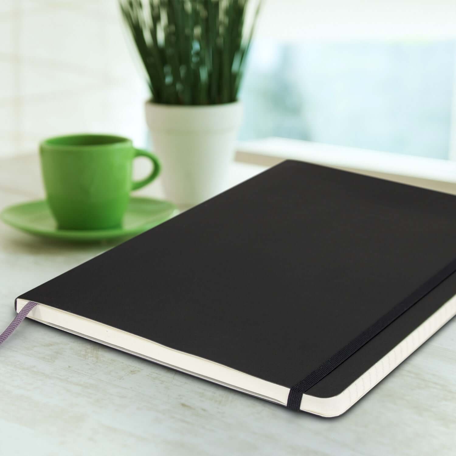 Moleskine Classic Soft Cover Notebook - Extra Large placed on a table with a green mug and plant.