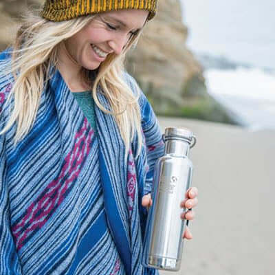Woman smiling while holding a Klean Kanteen Insulated Reflect Stainless Steel Bottle on a beach.