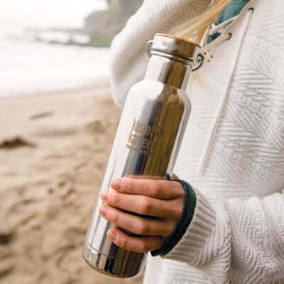 Klean Kanteen Insulated Reflect Stainless Steel Bottle held at the beach, showcasing its eco-friendly design.