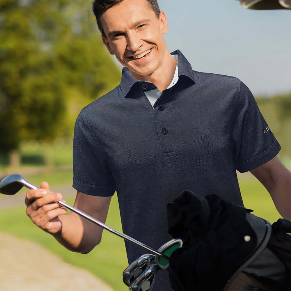 A smiling man wearing a Callaway Oxford Pique Polo Shirt, holding a golf club, enjoying outdoor activities.
