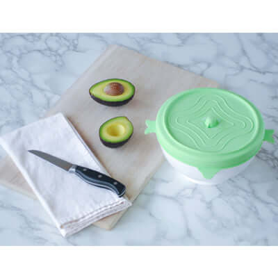 Storage Lid Set with green silicone lid, avocado halves, knife, and cloth on marble countertop.