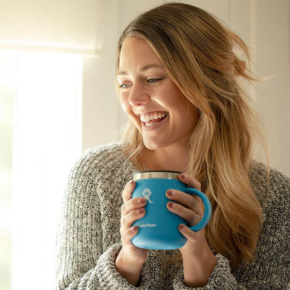 Woman smiling while holding a HydroFlask 12oz Coffee Mug, enjoying her drink in a cozy setting.
