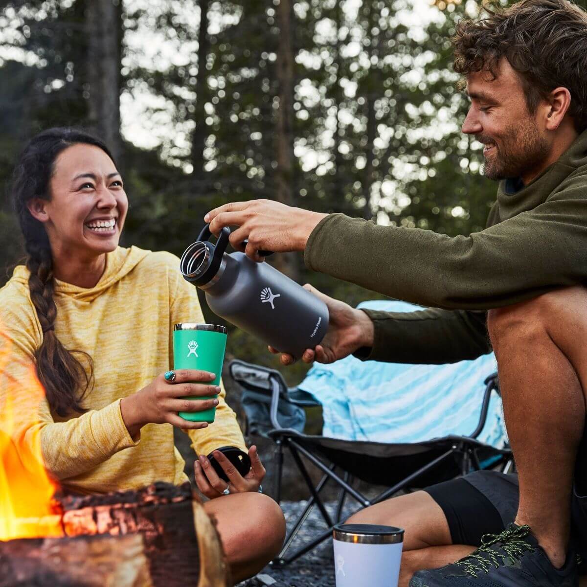 Two friends enjoying the outdoors with a HydroFlask 64 oz Growler Flask by the campfire, perfect for corporate gifts.